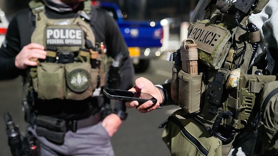 Federal officers in police tactical vests check a phone during an ICE Boston enforcement operation