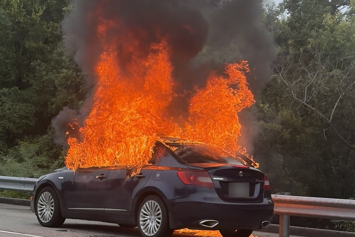 A dark sedan engulfed in flames beside a guardrail